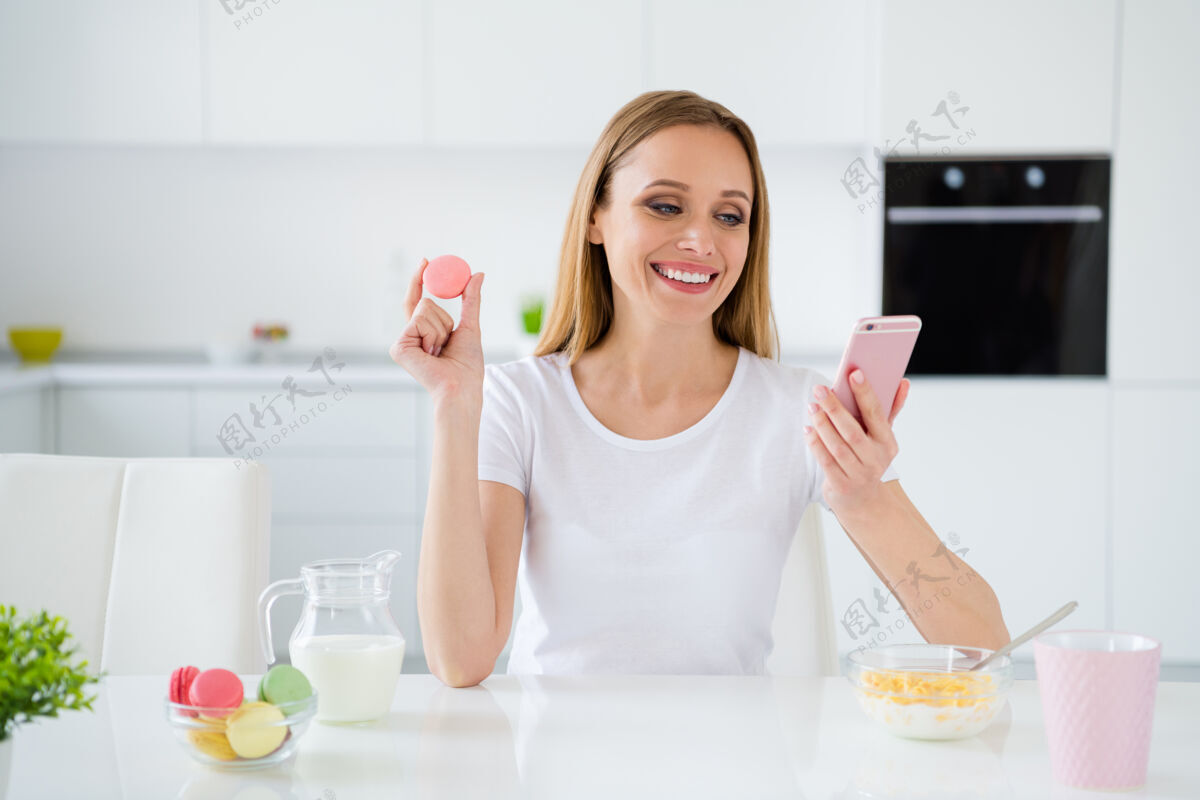 女人照片中漂亮的家庭主妇拿着电话和朋友聊天 桌上吃着五颜六色的杏仁饼格兰诺拉麦片早餐牛奶室内白光厨房甜点在线杯子