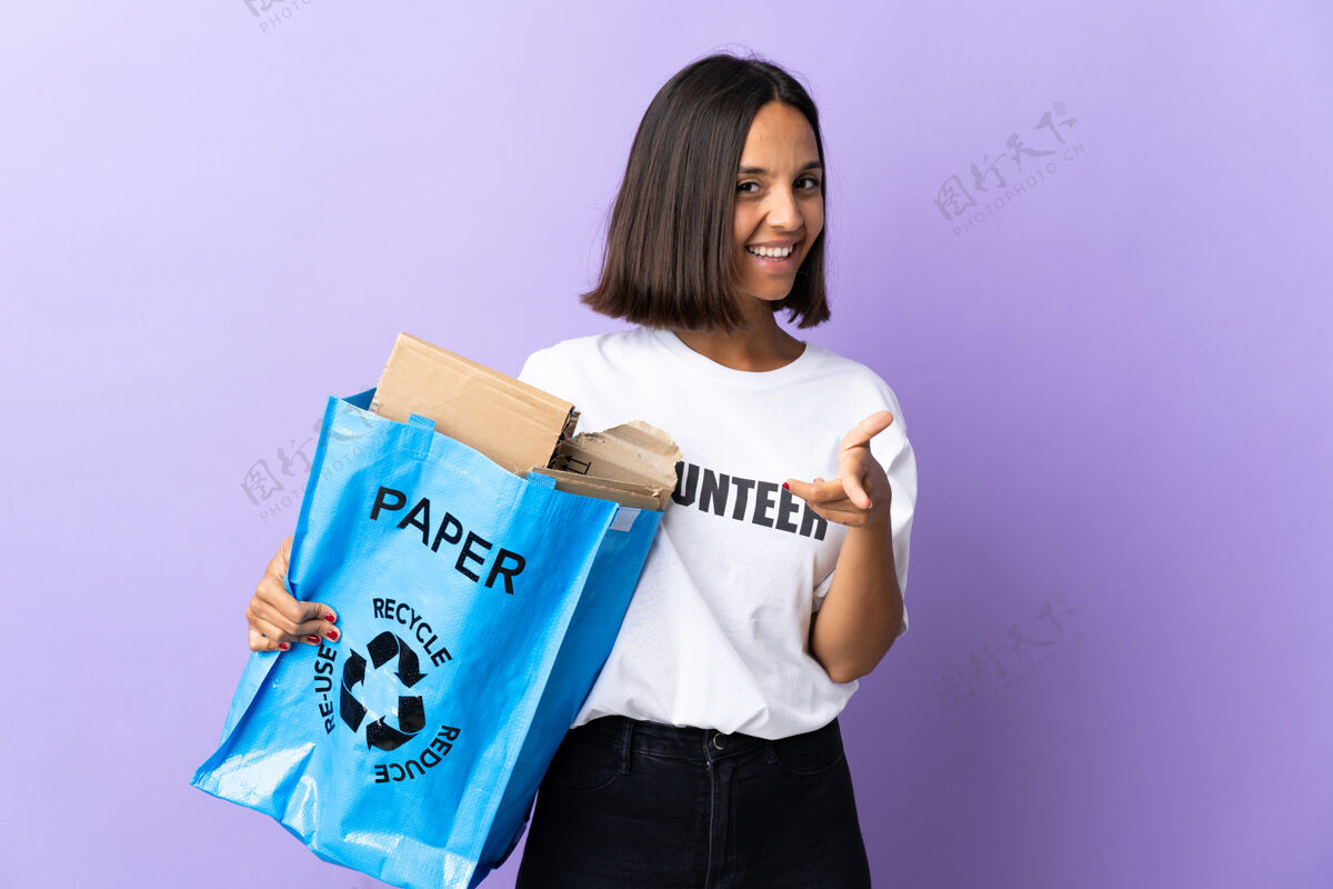 黑发年轻的拉丁女子拿着一个装满废纸的回收袋 孤立地指着紫色的正面微笑着保护重用手指