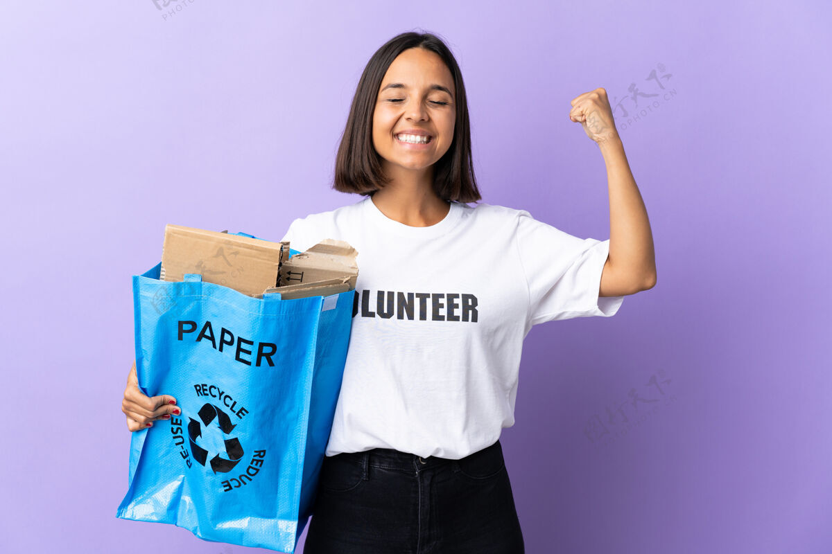姿态年轻的拉丁女子拿着一个装满废纸的回收袋 在紫色上做着孤军奋战的手势保护垃圾环境