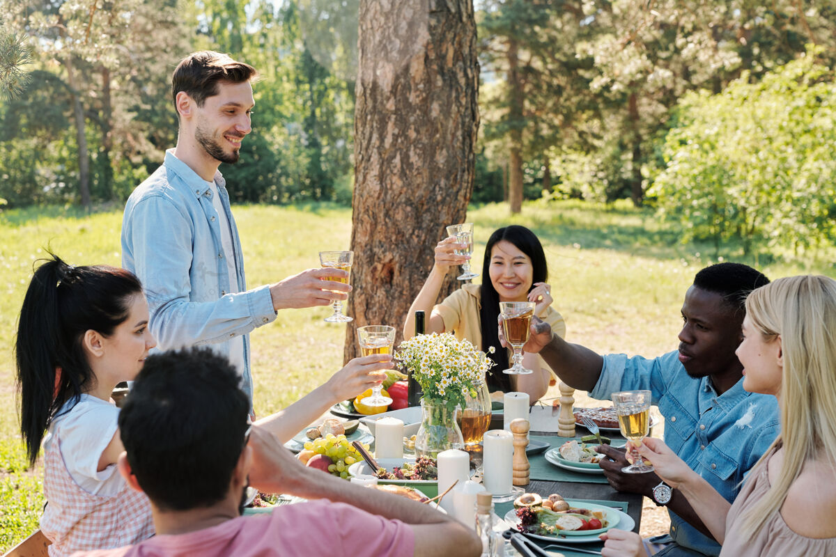 庆祝祝酒几位心平气和的国际友人将在松树下的户外晚宴上 一边在提供的节日餐桌上敬酒 一边碰杯欢呼快乐夏天