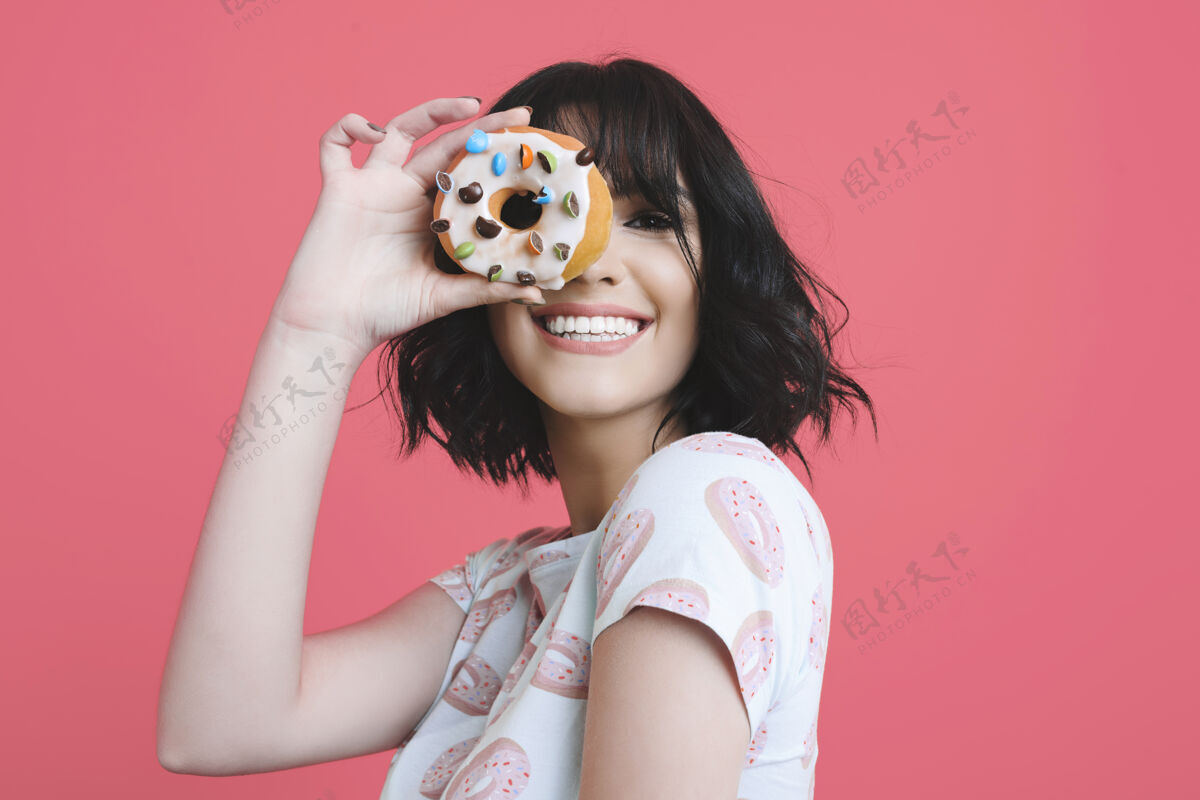 年轻一个可爱迷人的年轻白种女人穿着一件有甜甜圈的衬衫 透过一个甜甜圈 在粉红色的工作室背景下大笑化妆爱情站着
