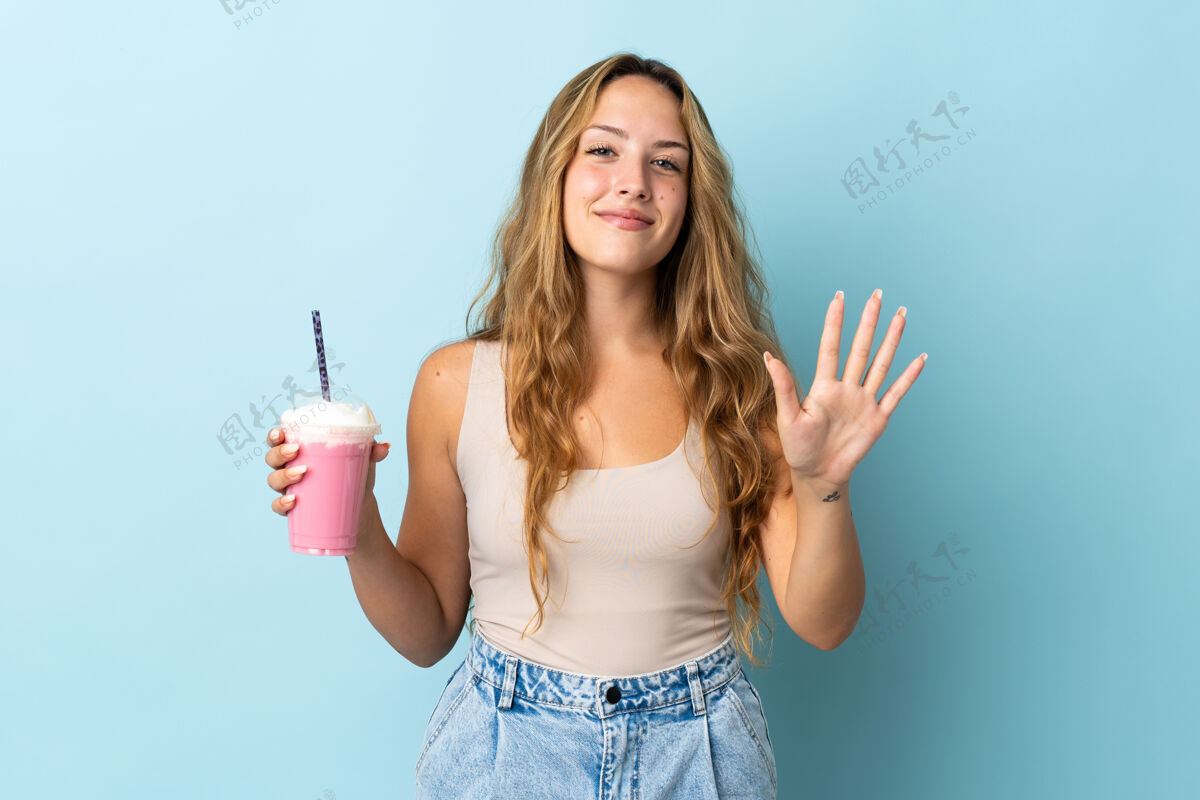 女士拿着草莓奶昔的年轻女子被隔离在蓝色的墙上 用手指数着五个奶昔杯子女孩