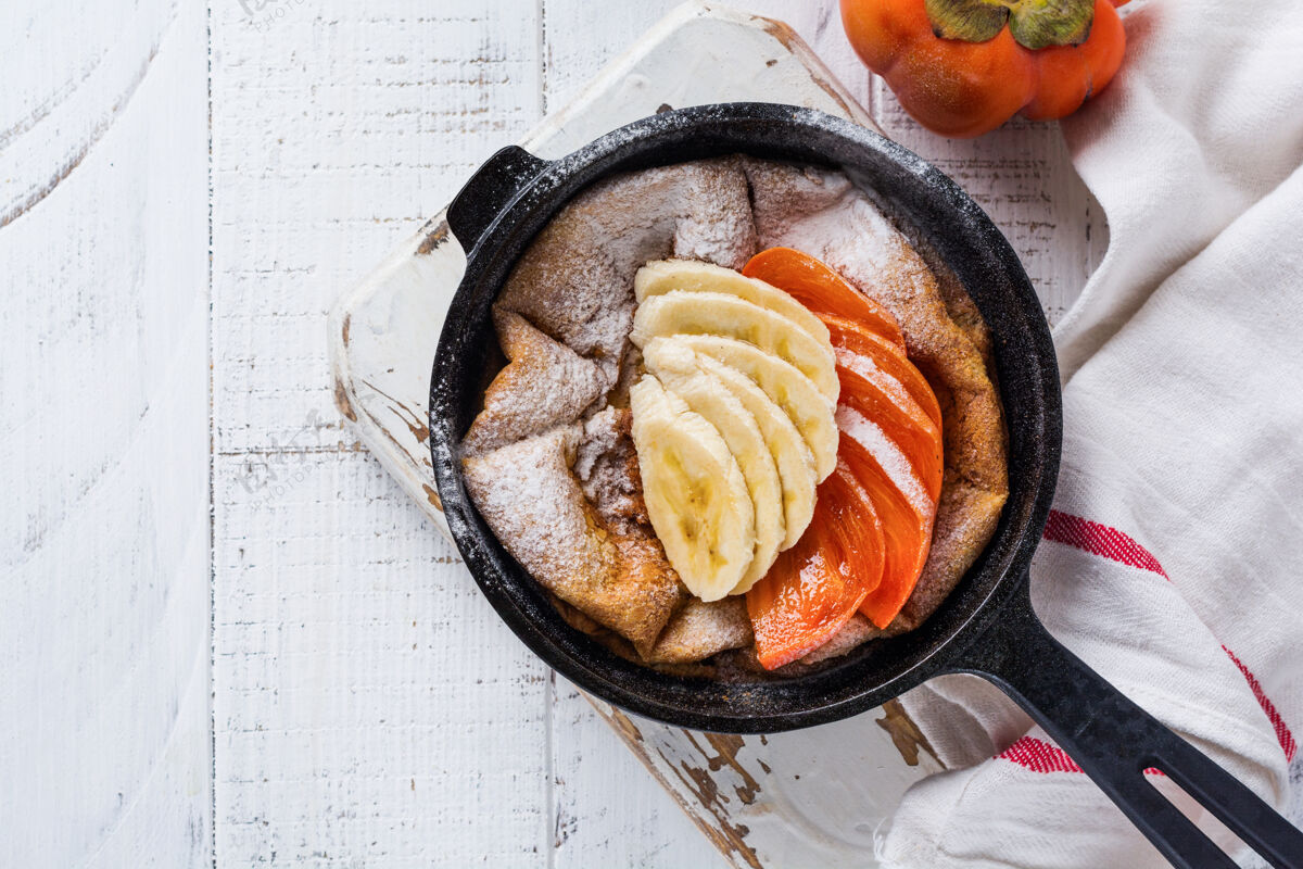 美味荷兰宝宝煎饼用苹果 柿子 香蕉 肉桂放在小铁锅的木制台面上早上派柿子
