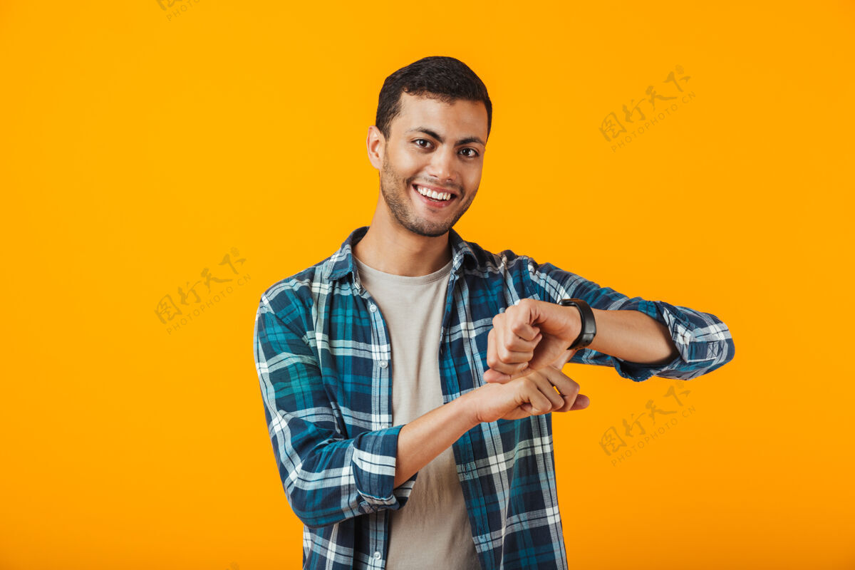 英俊一个穿着格子衬衫的年轻人孤零零地站在橙色的墙上 看着他的手表迟到人自信