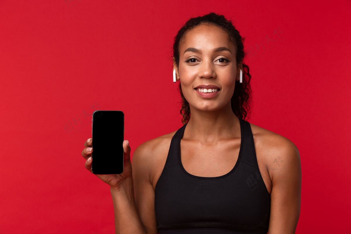 教练身着黑色运动服 使用智能手机和无线耳机 隔离在红墙上的女性非洲裔美国妇女的肖像女性耳塞健身房