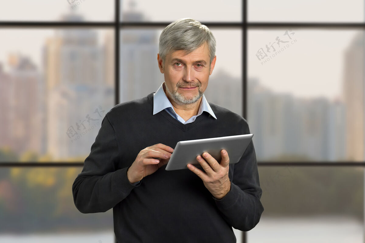 男人带着电脑平板电脑的成熟男人的肖像严肃的白发男人拿着电脑平板电脑在办公室的窗户上特写窗口严肃个人