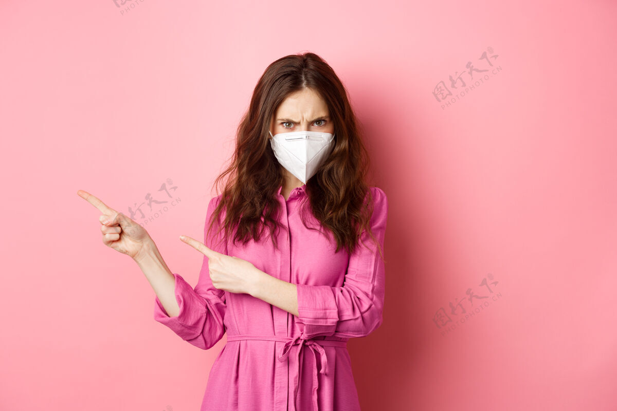 社交距离科维德-19 隔离和社会距离概念愤怒的女人皱着眉头 看起来很有判断力 戴着口罩 指着左边的手指 粉色的墙壁女孩女性口罩