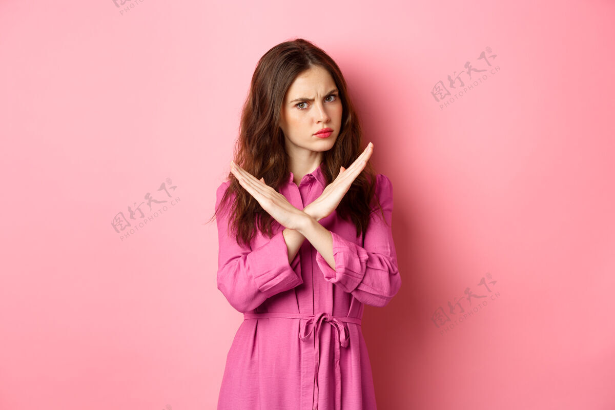 候选人不高兴的愤怒的女人挡住了提议 做了一个交叉停车的手势 说不 摇头否定回答 站在粉红色的墙上手势否认停止