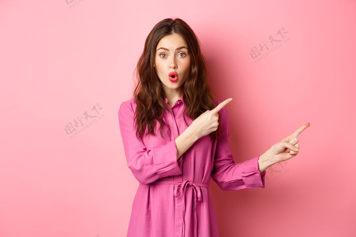 女孩美丽和时尚惊讶的女人手指指向右边 展示广告在旁边的空间 站在粉红色的墙壁快乐积极惊人