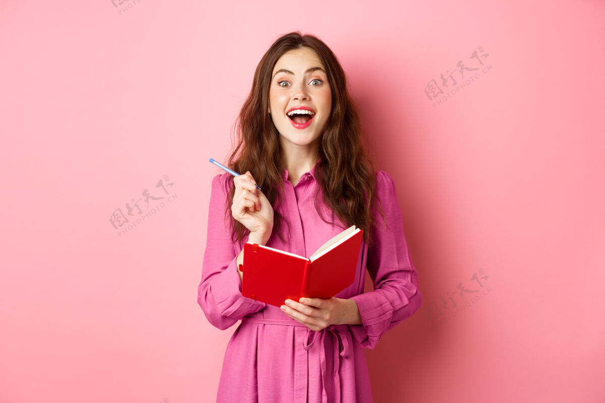 购物兴奋的年轻女子有很好的想法 把她的想法写在计划者 拿着笔记本日记 微笑着惊讶 站在粉红色的墙上笔记本快乐女孩
