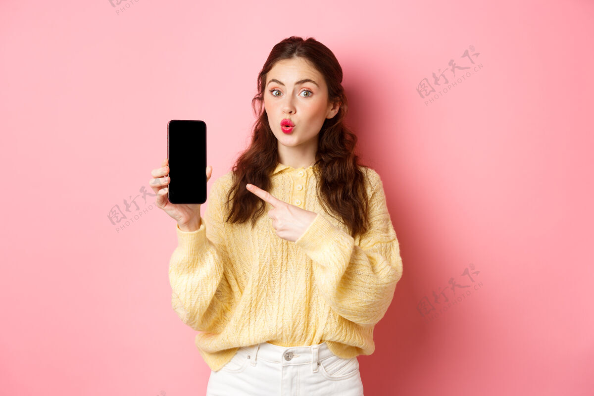 应用程序科技和网上购物兴奋的女孩看起来很好奇 手指指着空手机屏幕 在智能手机上显示应用程序 站在粉红色的墙上应用程序魅力黑发