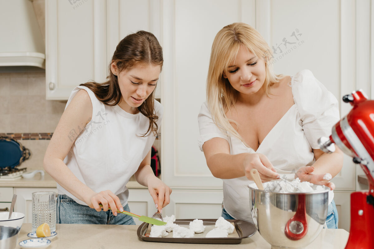 白人在厨房里 两个微笑的年轻女人正在用勺子和肩胛骨把搅打好的蛋白酥皮放在托盘上金发鸡蛋女人