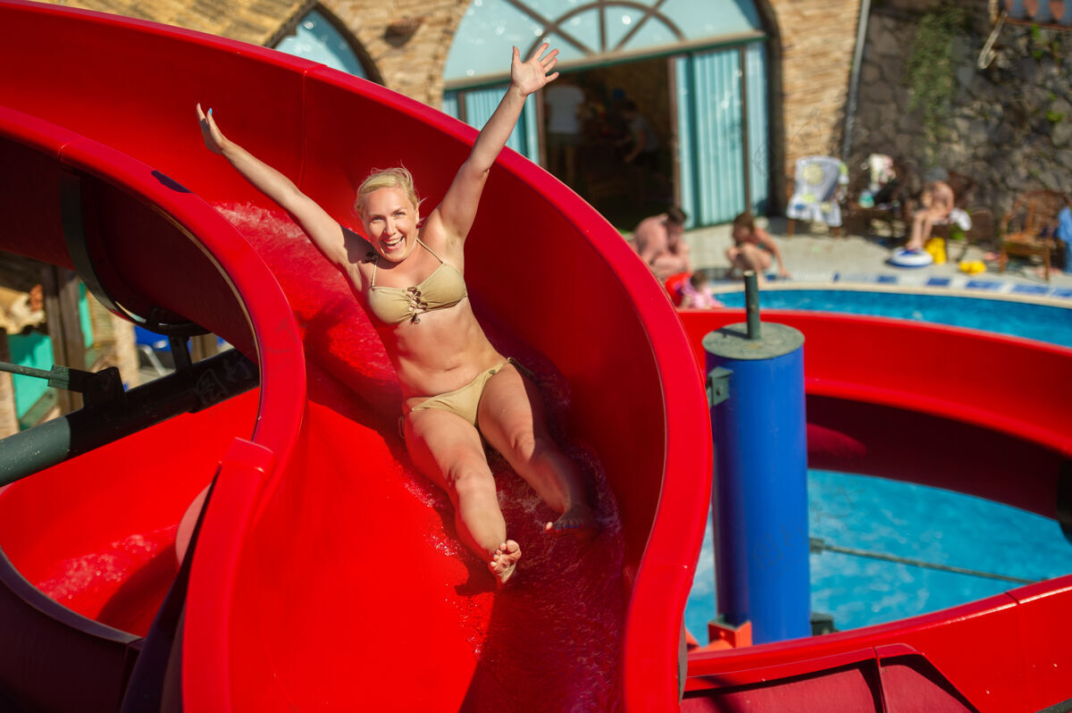 塑料度假期间 一个穿着泳装的快乐女人从水上公园的红色滑梯上下来欢呼比基尼滑梯