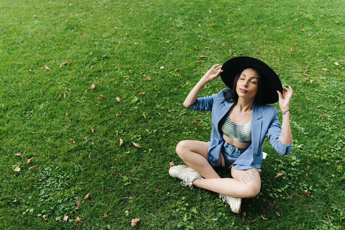 草地穿着蓝色条纹夹克和黑色夹克的年轻女子的时尚肖像帽子女人坐在城市公园的绿色草坪上休闲绿草模特