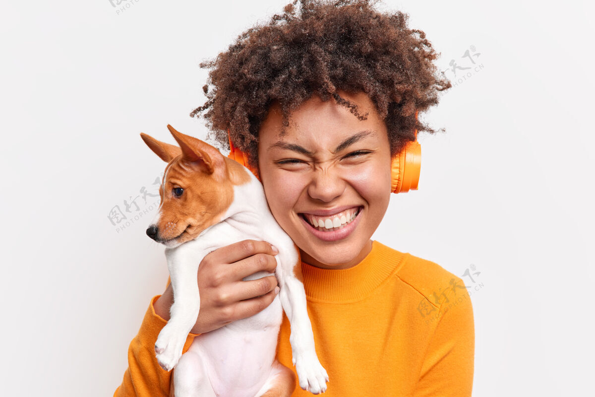 拥抱人与动物的友谊概念喜出望外的卷发女子微笑温柔地抱着小纯种小狗紧挨着脸 隔着白墙隔着无线耳机听音乐动物欣喜若狂狗