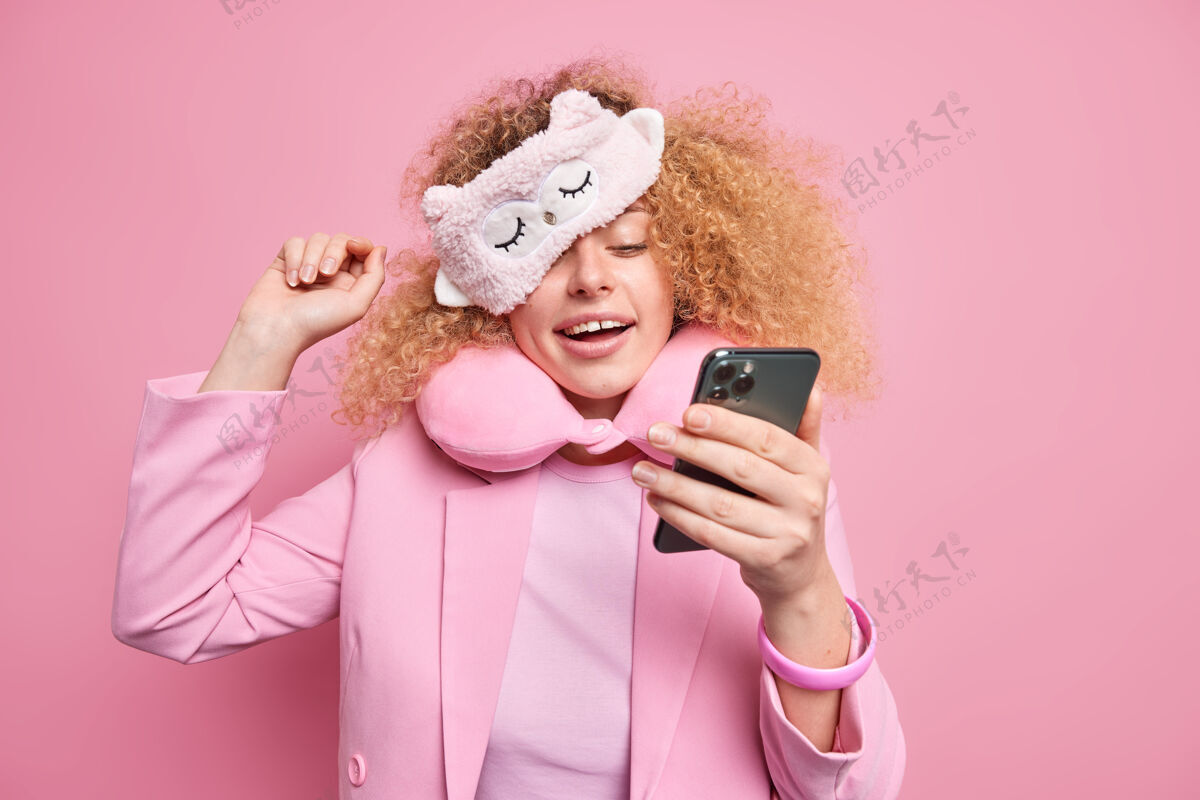 姿势积极无忧无虑的女人沉迷于现代科技 醒来后通过智能手机查看社交网络的新闻 戴着柔软的睡眠面罩 穿着正式的衣服 在粉色的墙上聊天漂亮女人放松