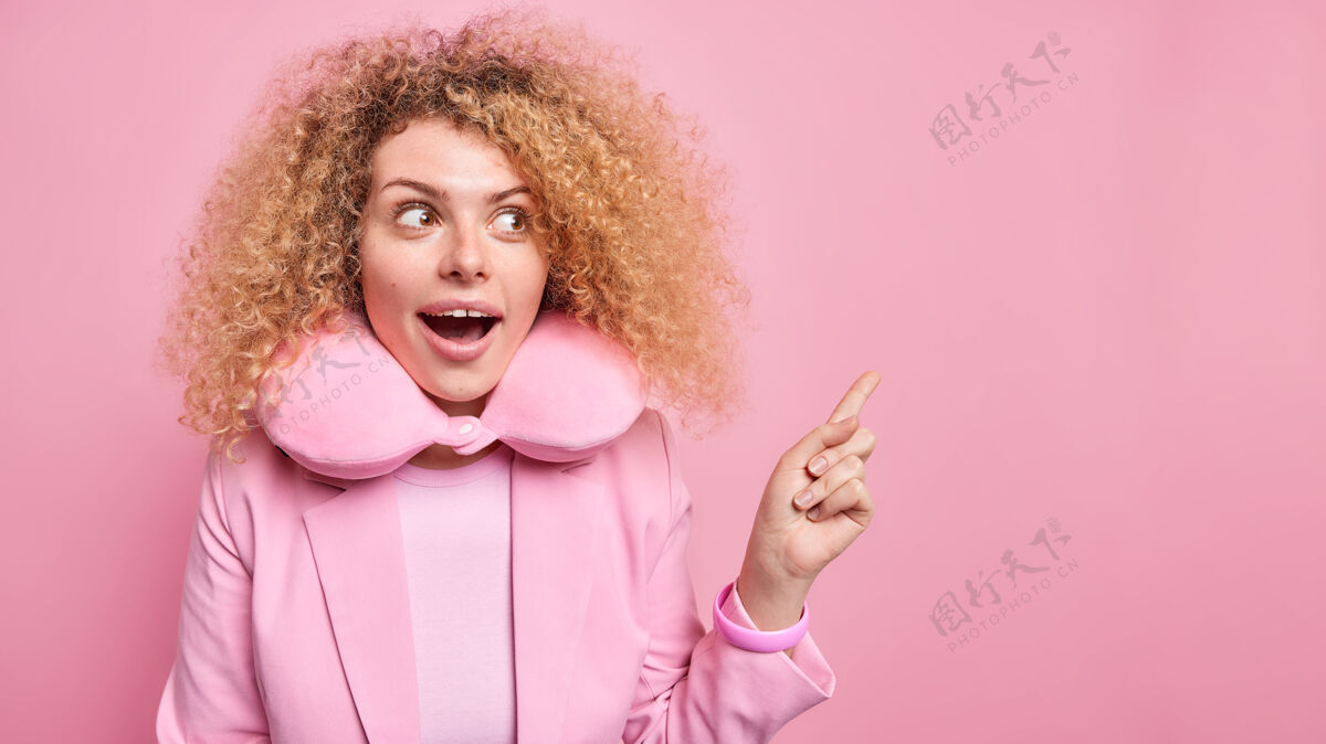 女性迷人惊讶的女人从惊奇中张嘴有卷曲浓密的头发惊讶地表示在空白处佩戴舒适的颈枕展示产品粉色墙壁单色年轻夹克
