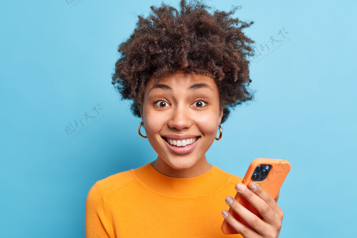 非洲一位留着卷发 面带微笑的年轻女子的肖像使用手机在线聊天下载新应用程序looksgladfully身穿橙色套头衫隔着蓝色的墙壁欢乐乐观欢呼