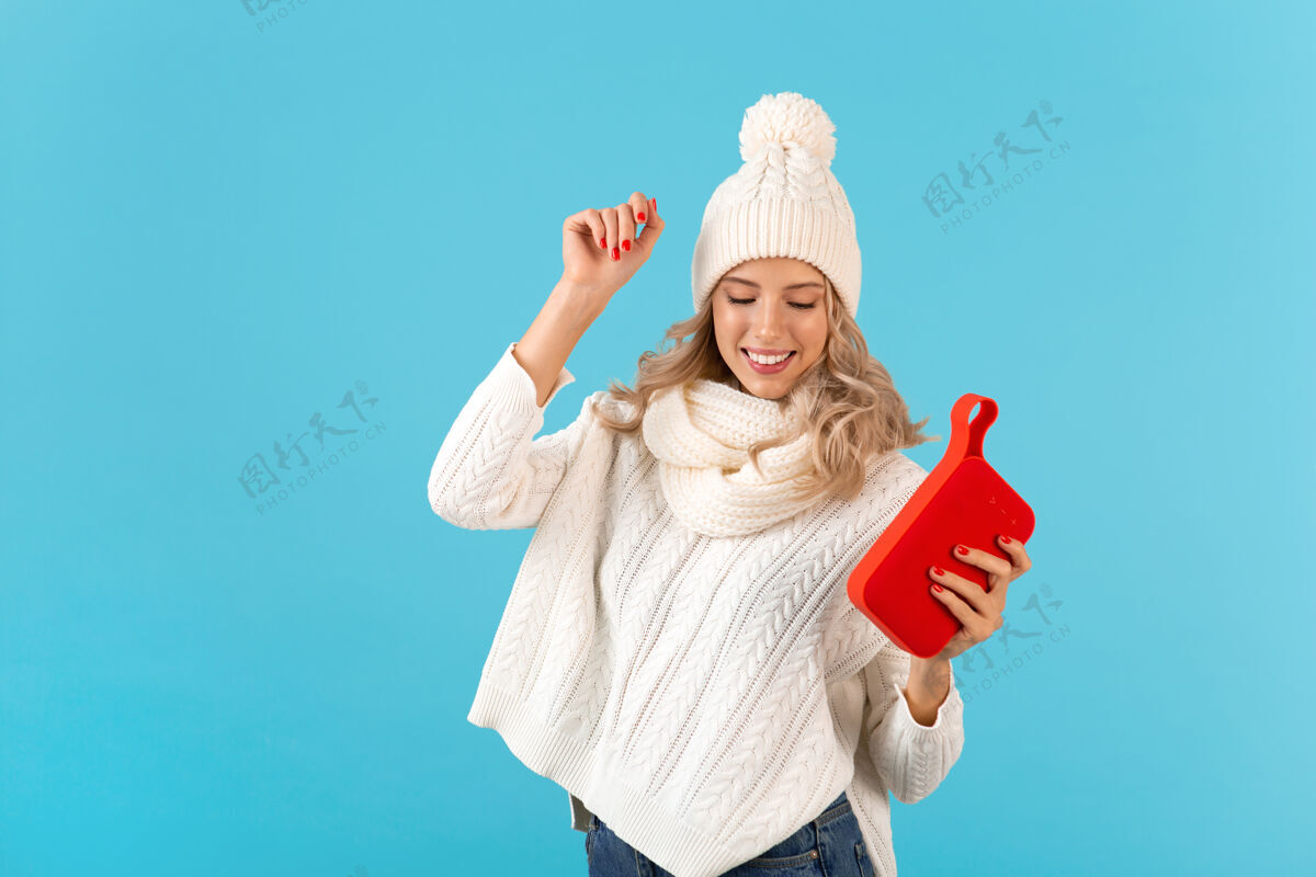 季节时尚的金发微笑美丽的年轻女子手持无线扬声器听音乐快乐跳舞穿着白色毛衣和针织帽子冬季时尚摆造型温暖女人音频