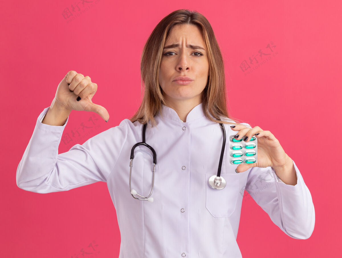 年轻人年轻的女医生穿着医用长袍 手持听诊器 拇指朝下 被隔离在粉红色的墙上药片按住感觉