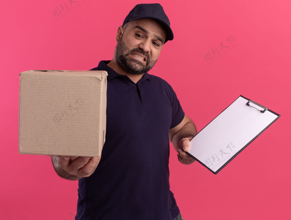 人一个穿着制服 戴着帽子的中年送货员拿着剪贴板 拿着盒子对着隔离在粉红色墙上的摄像机剪贴板盒子人