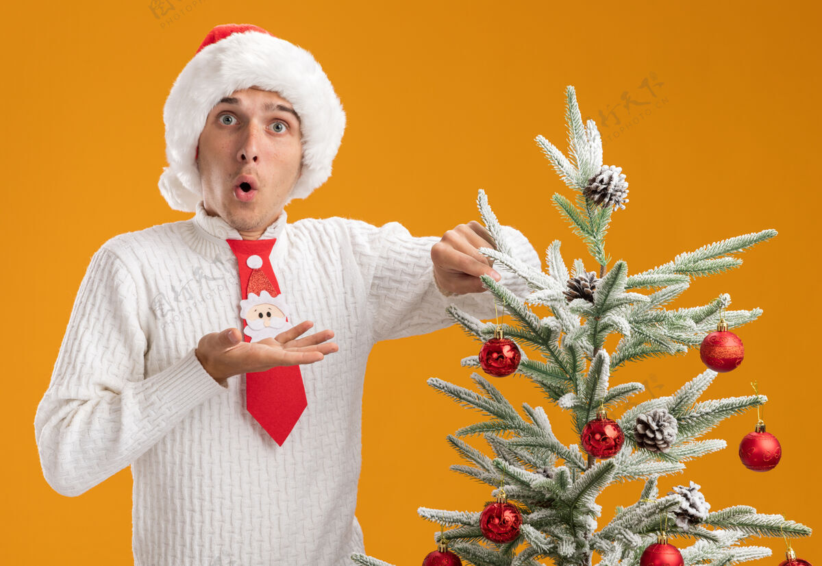 小伙子令人印象深刻的年轻帅哥戴着圣诞帽和圣诞老人领带站在圣诞树旁装饰圣诞球装饰看起来空手孤立在橙色的墙上近空帽子