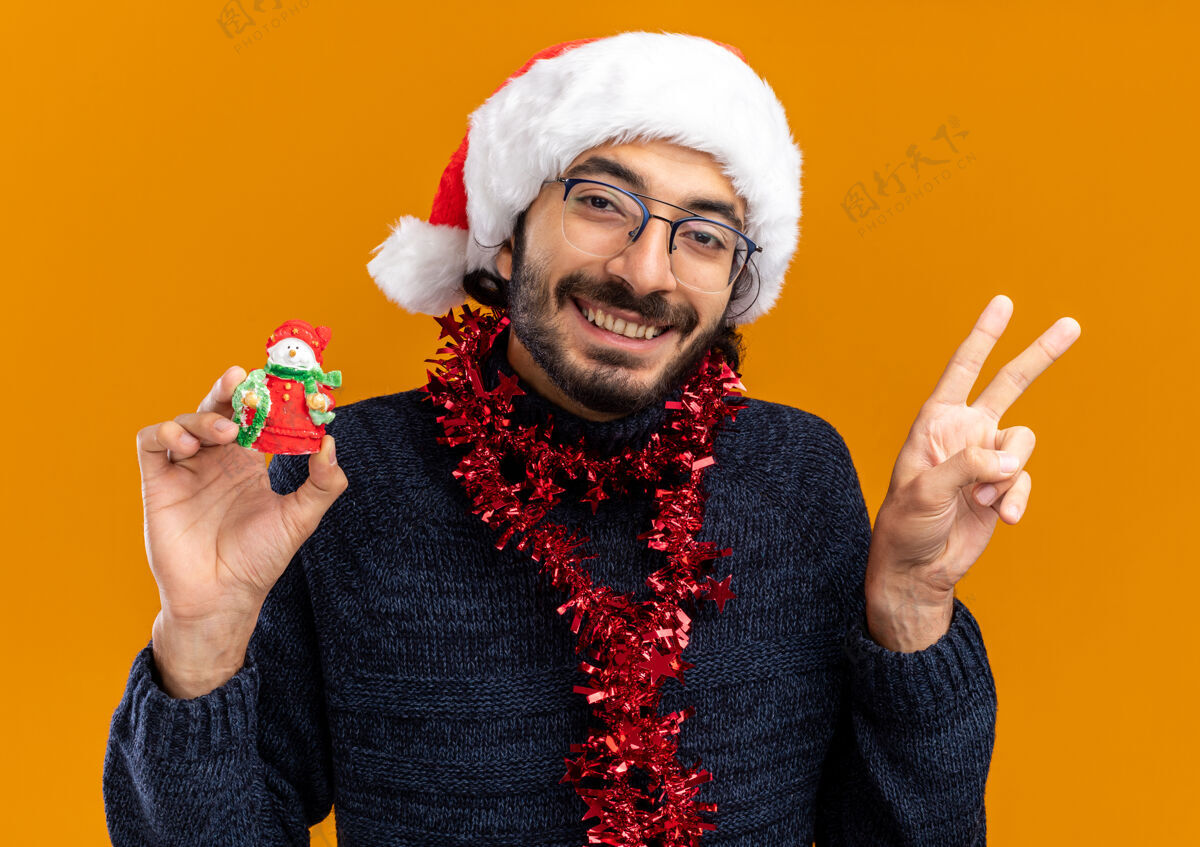 男子微笑着的年轻帅哥戴着圣诞帽 脖子上戴着花环 手里拿着玩具 在橙色的墙上展示着与世隔绝的和平姿态脖子显示和平
