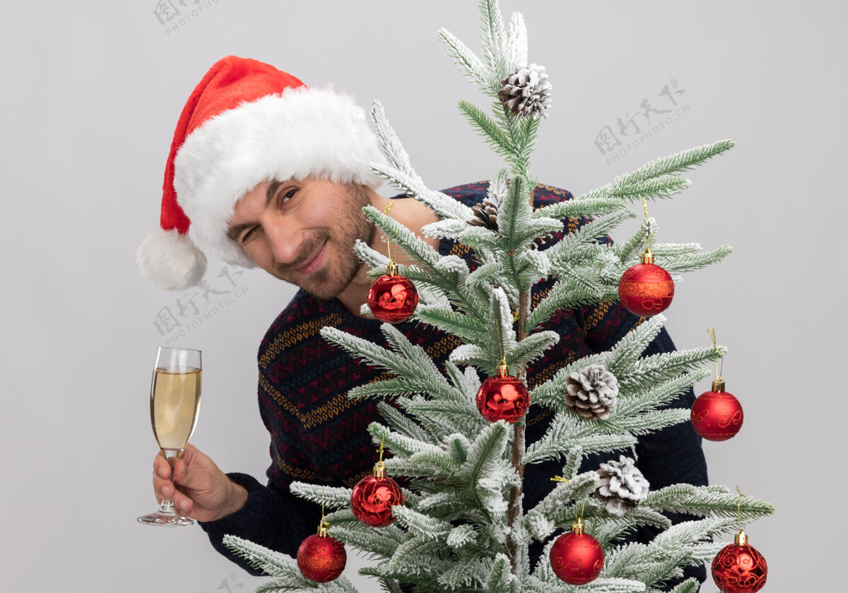 帽子高兴的年轻白人男子戴着圣诞帽站在圣诞树后面望着拿着一杯香槟在白色的墙上眨眼香槟戴着白种人