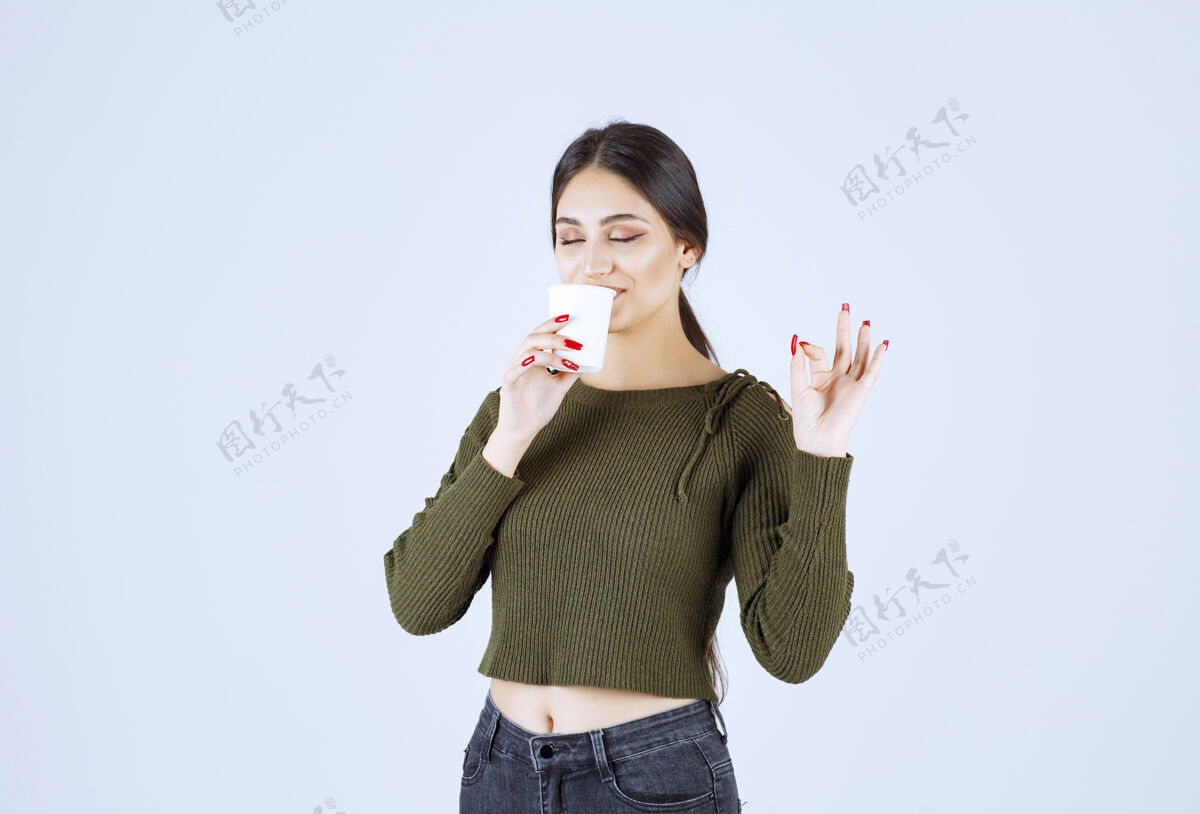 成人年轻的黑发女人喝着一杯塑料茶 并发出“好”的信号黑发女孩杯子