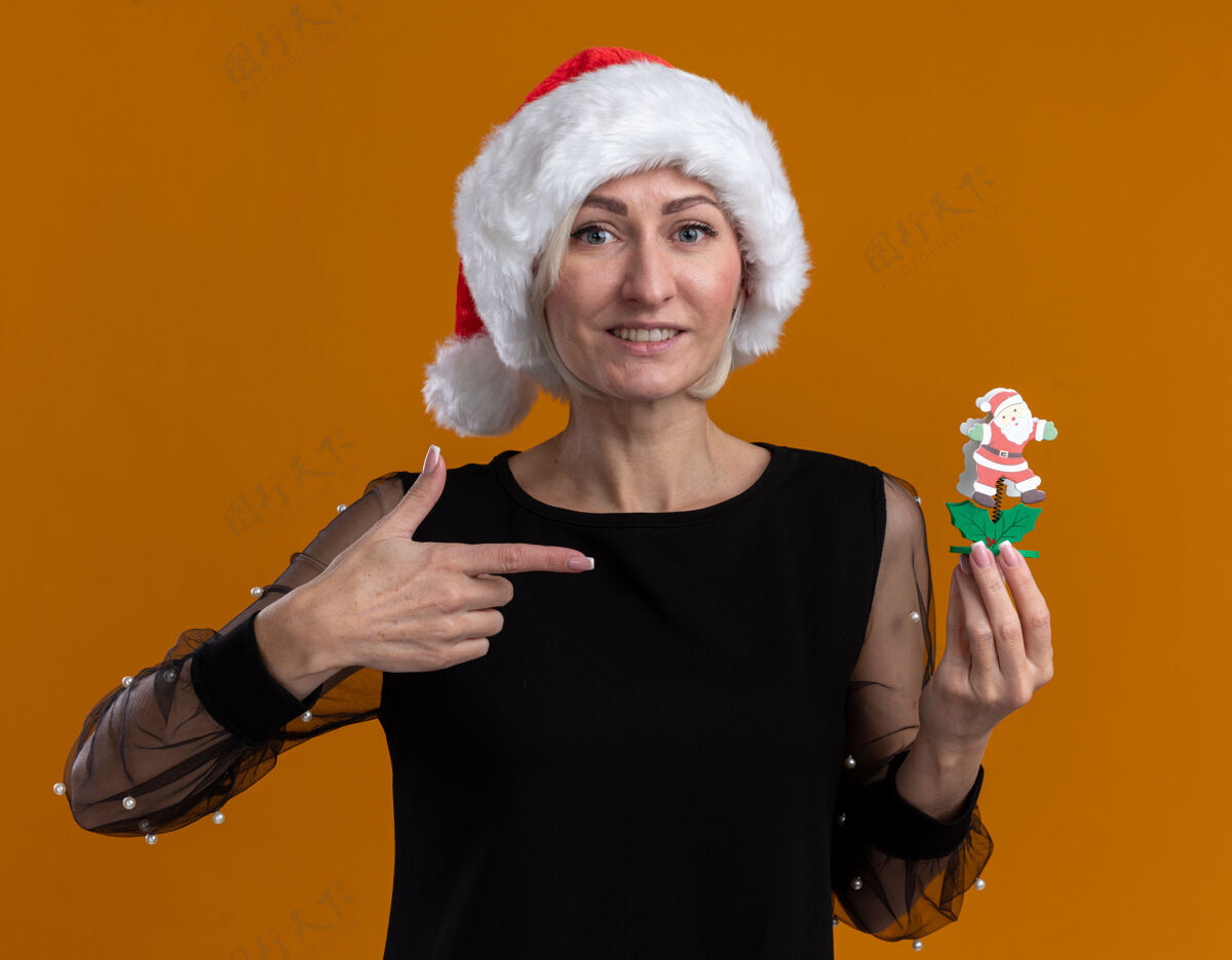 金发微笑着的中年金发女人戴着圣诞帽看着拿着并指着隔离在橙色墙上的圣诞老人玩具指着帽子女人