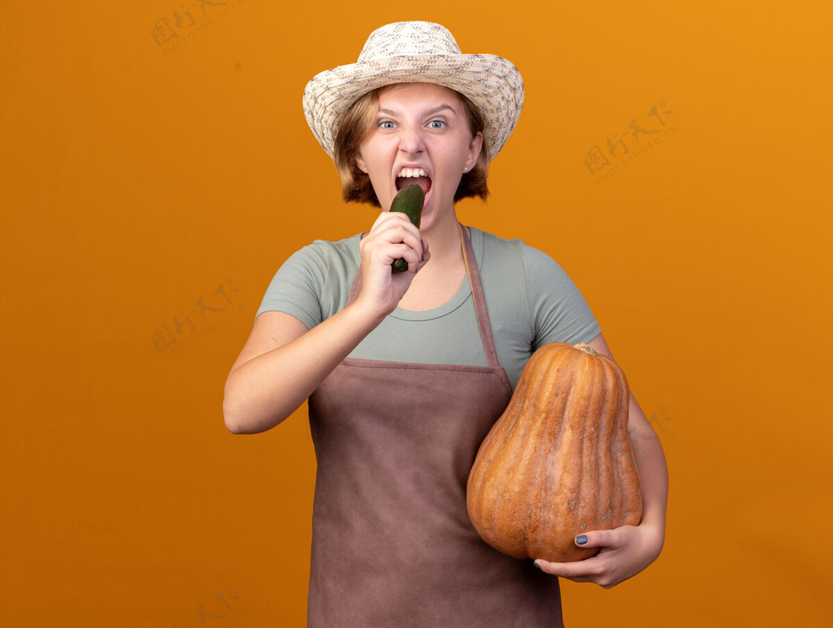 帽子饥饿的年轻斯拉夫女园丁戴着园艺帽 手里拿着南瓜 咬着黄瓜花园橘子南瓜