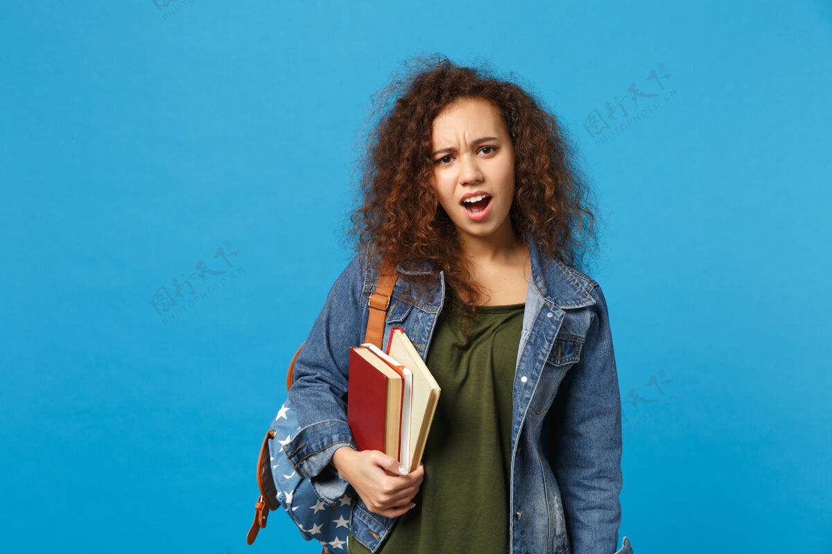 学习穿着牛仔服 背着书包的悲伤的年轻学生把书孤立地放在蓝色的墙上截止日期知识青少年