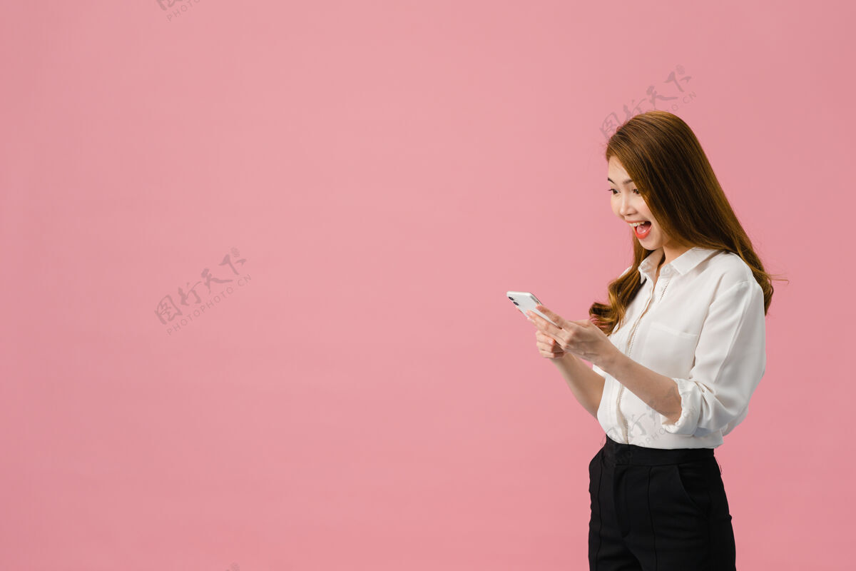 媒体年轻的亚洲女士用手机 表情积极 笑容灿烂 穿着休闲服 感觉幸福 孤立地站在粉色背景上商务女性亚洲人商业