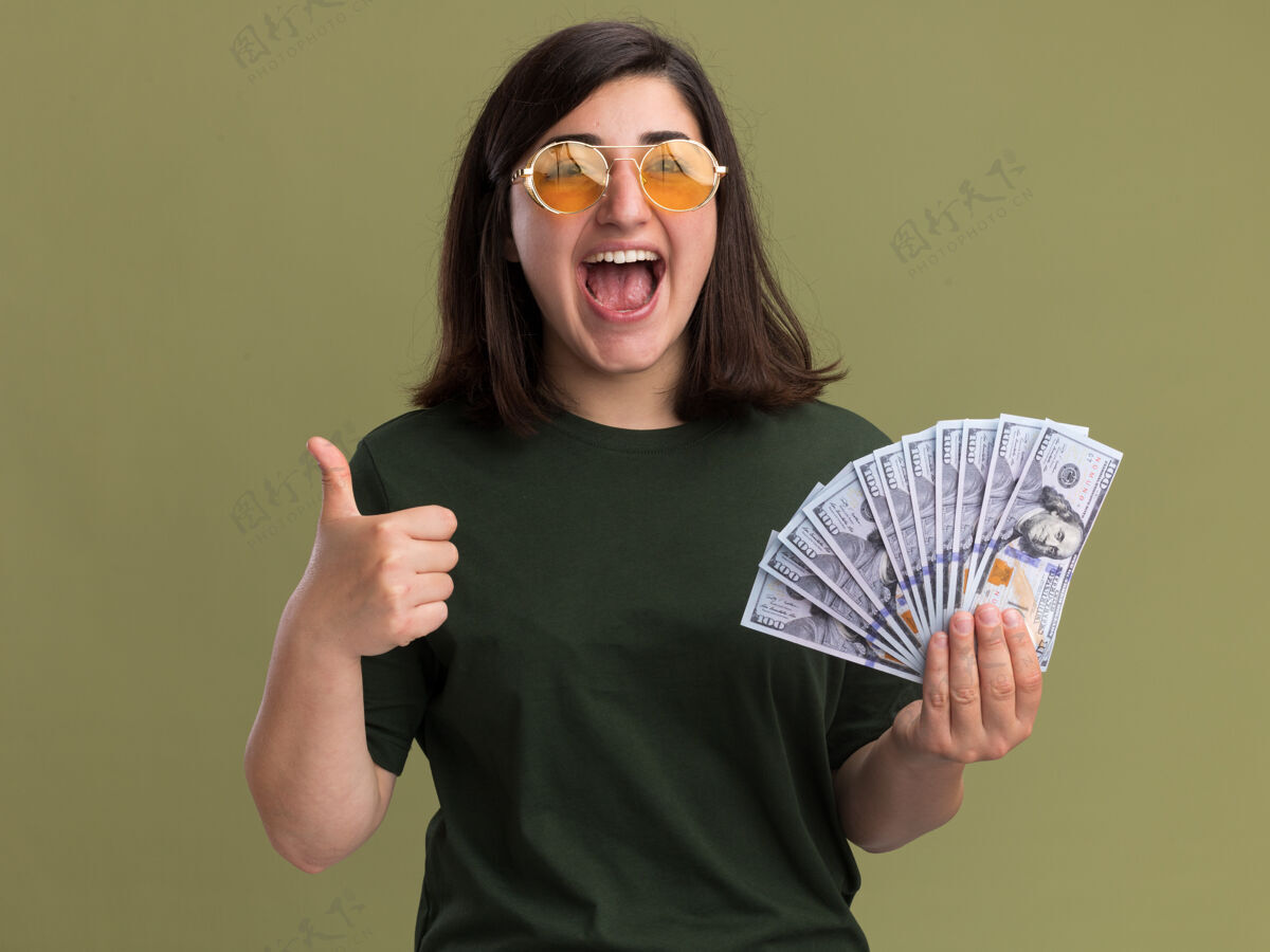 年轻戴着太阳眼镜的年轻漂亮的白人女孩高兴地竖起大拇指 手里拿着钱太阳眼镜拇指