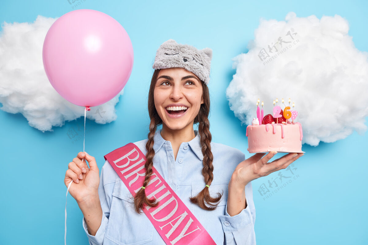 梦幻两个小辫子集中在上面的女孩表情欢快地拿着喜庆蛋糕和氦气球庆祝26岁生日在派对上等待朋友接受祝贺年轻人蛋糕睡眠面膜