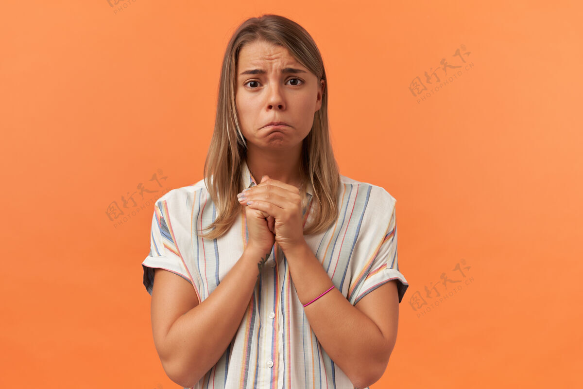 心烦一个穿着便服的悲伤而不快乐的年轻女子 双手放在祈祷的位置 看着被隔离在橙色墙壁上的前方美丽姿势模特