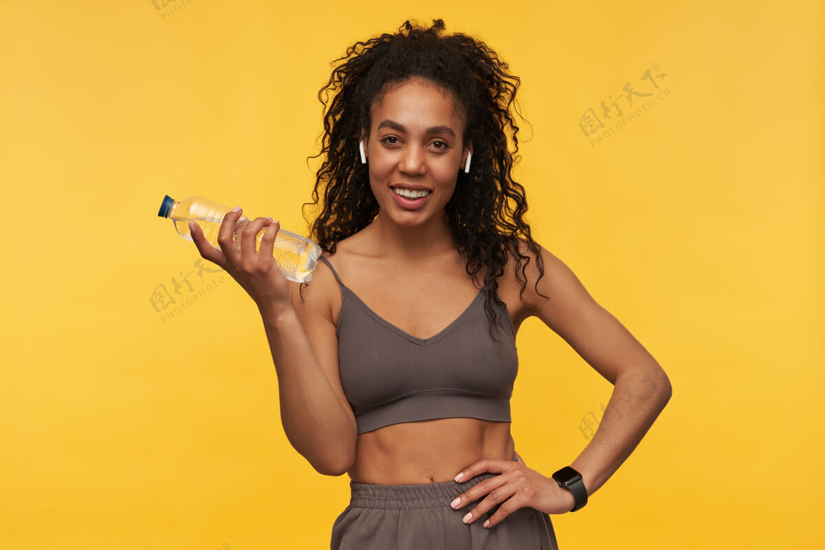 站立快乐迷人的年轻女运动员 戴着无线耳机和智能手表 站在黄色的墙上 手里拿着一瓶水人休闲女运动员