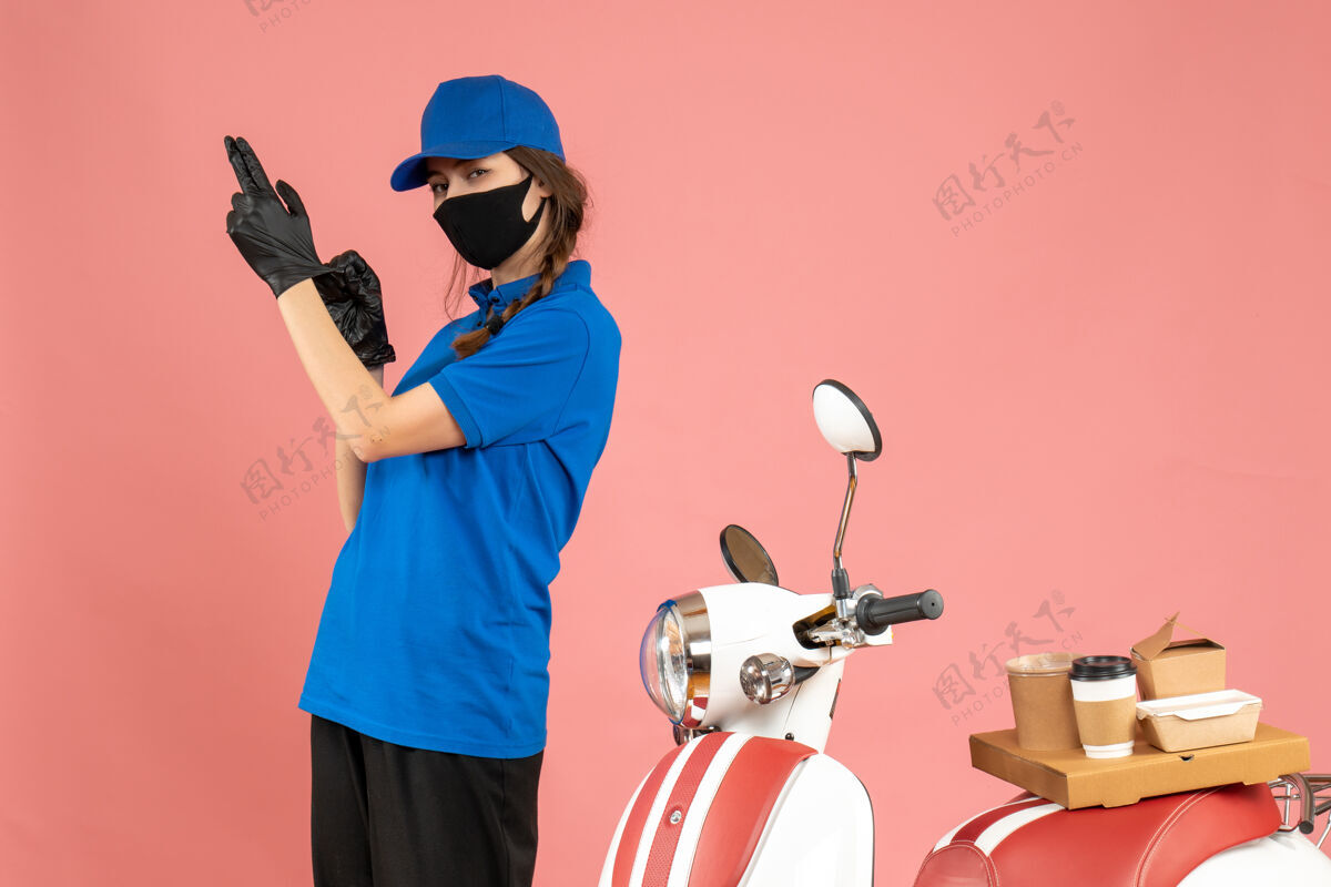 立场顶视图自信的信使女孩戴着医用口罩手套站在摩托车旁边 咖啡蛋糕在柔和的桃色背景上成人蛋糕高尔夫