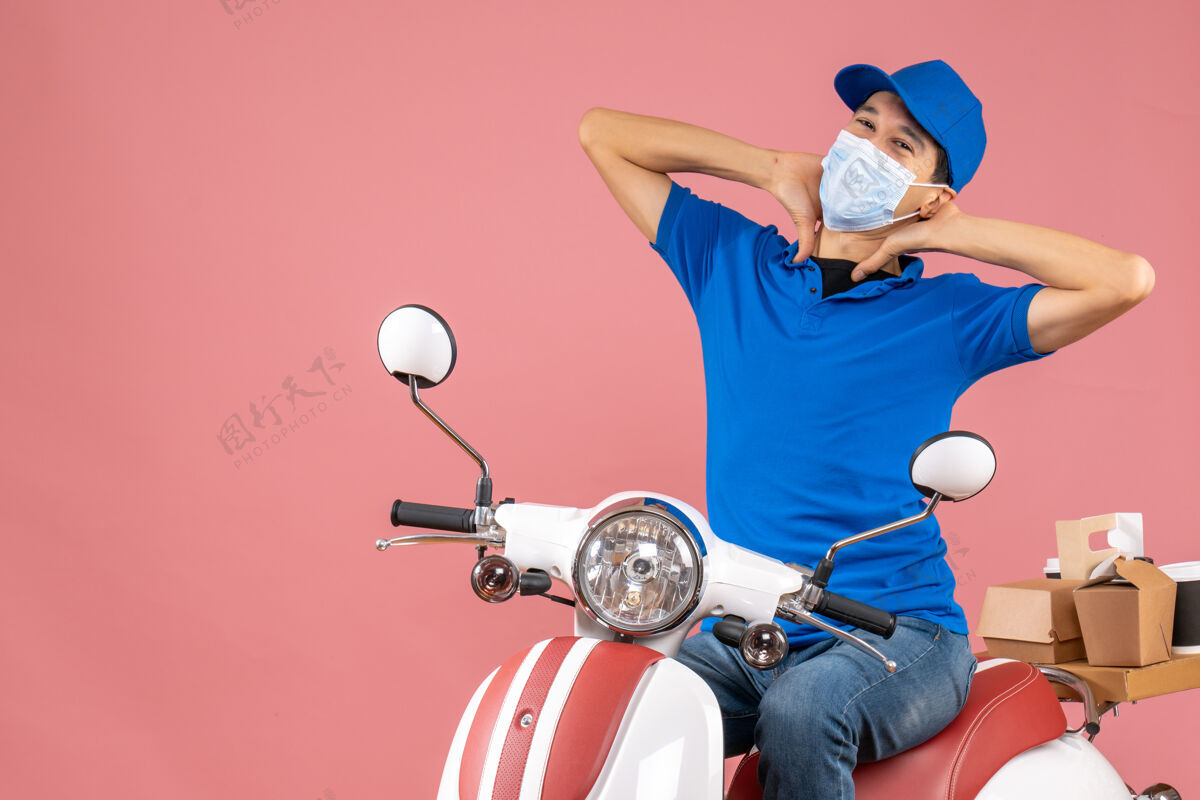 正面正面图：戴着医疗面罩戴着帽子的快递员坐在滑板车上 在柔和的桃色背景下传递订单摩托车粉彩人