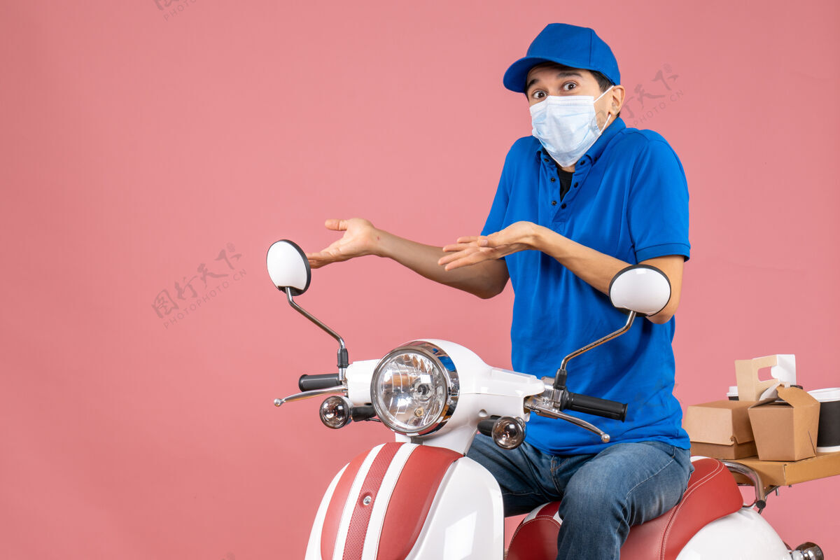 滑板车困惑的送货员戴着医用面罩 戴着帽子 坐在粉彩桃色背景的踏板车上人物坐粉彩