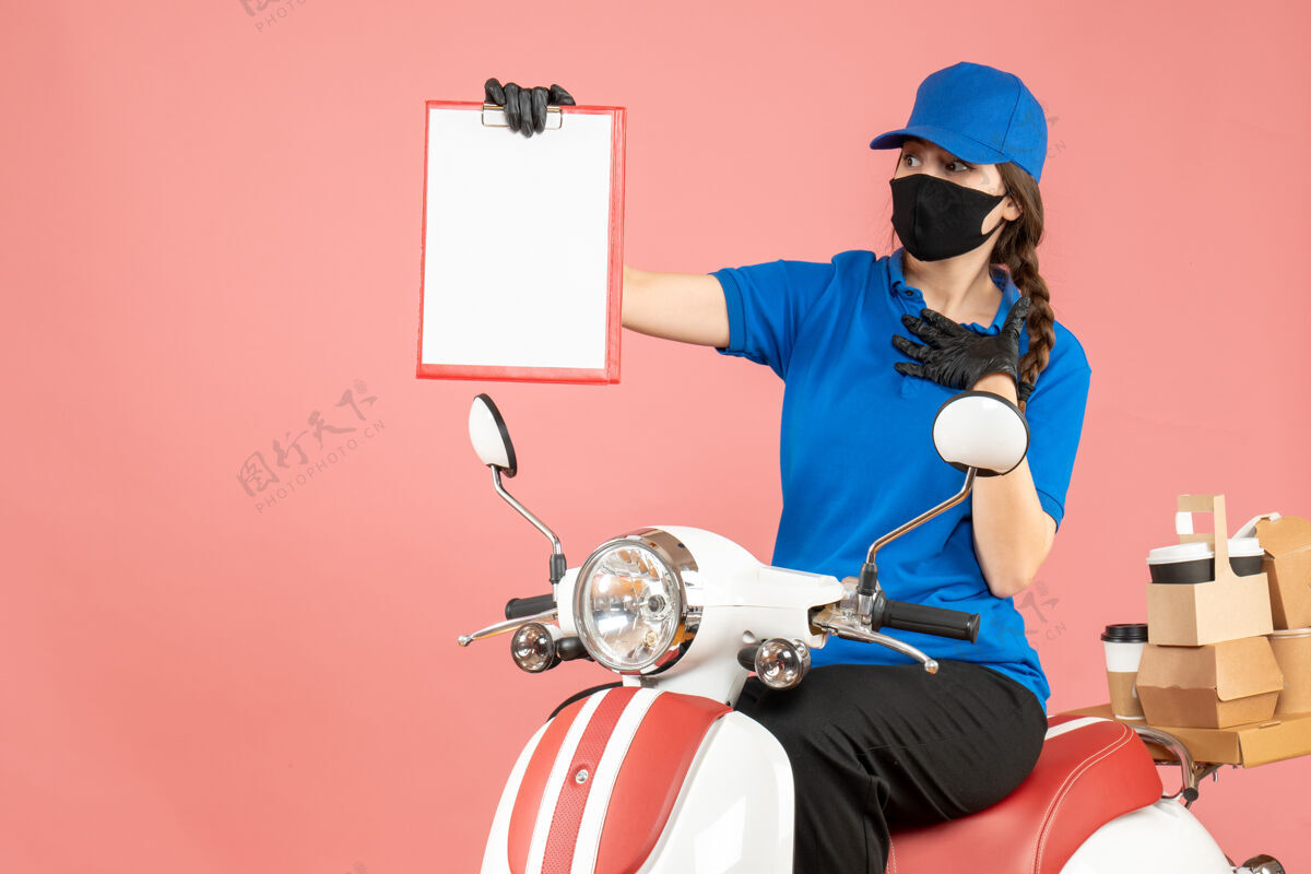 女信使困惑的女快递员戴着医用面罩和手套坐在滑板车上 拿着空纸 在粉彩桃色背景下传递订单空手套坐