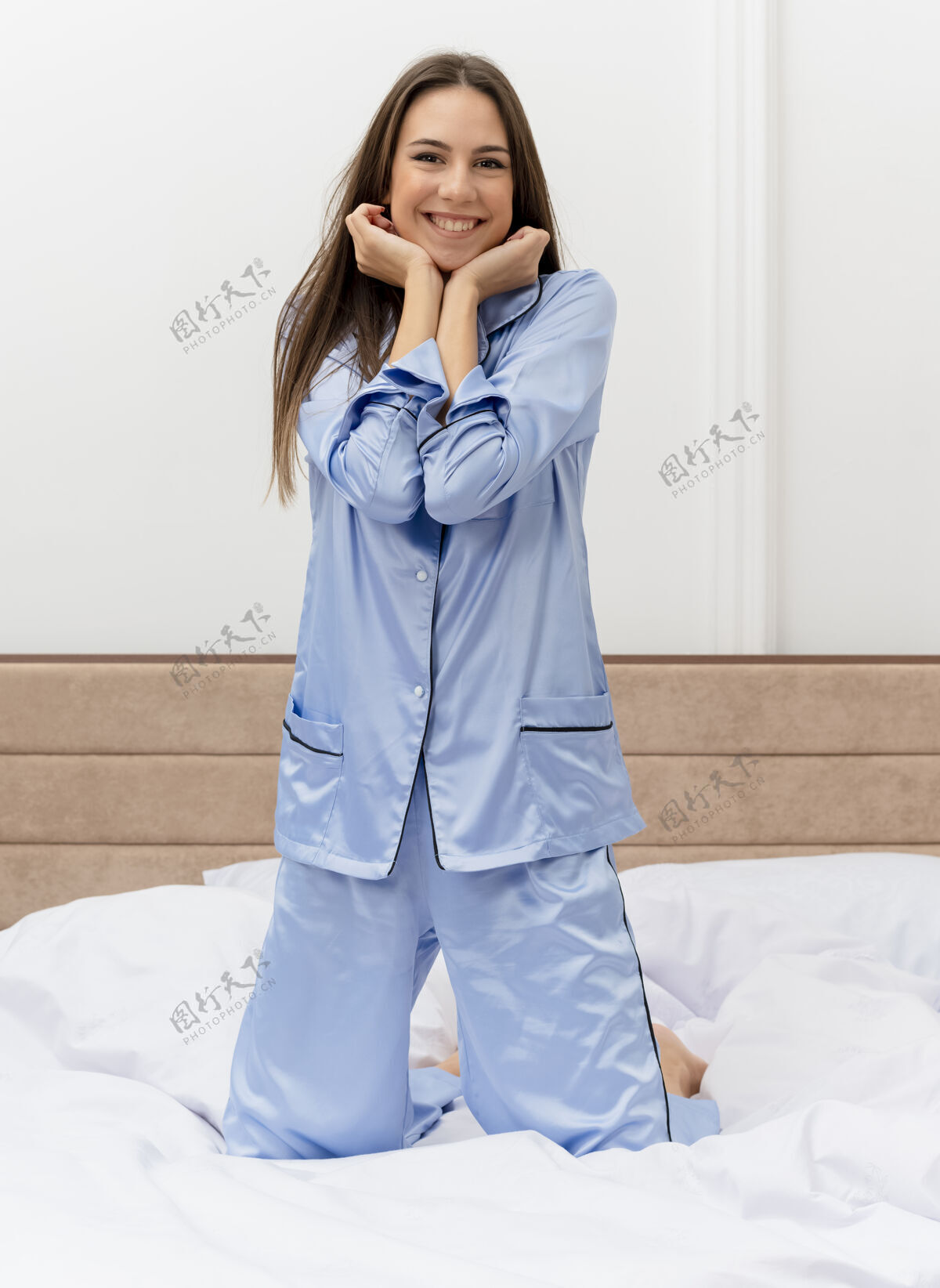 享受穿着蓝色睡衣的年轻美女坐在床上休息 享受周末的快乐和积极的微笑愉快地在卧室内部蓝色微笑美丽