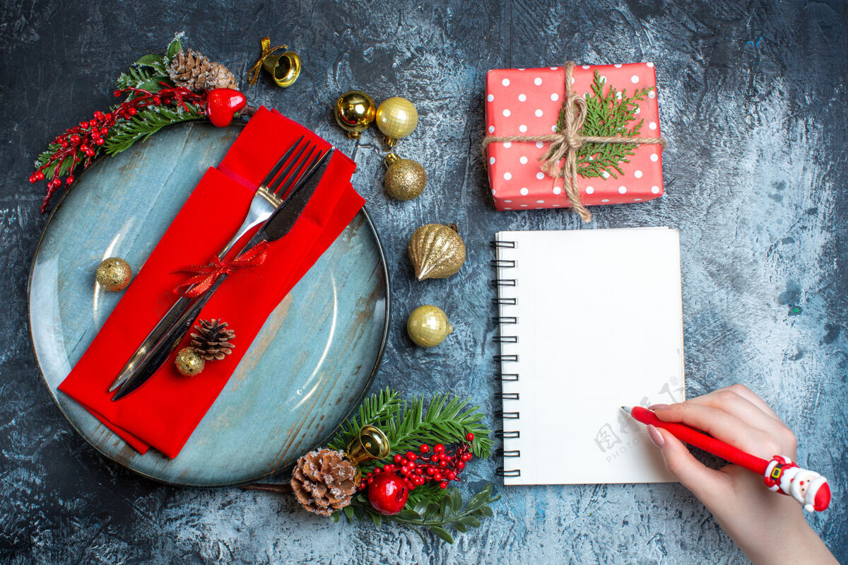 笔记本上图：手拿一支笔放在螺旋笔记本和餐具旁的礼品盒上背景餐具套装礼品