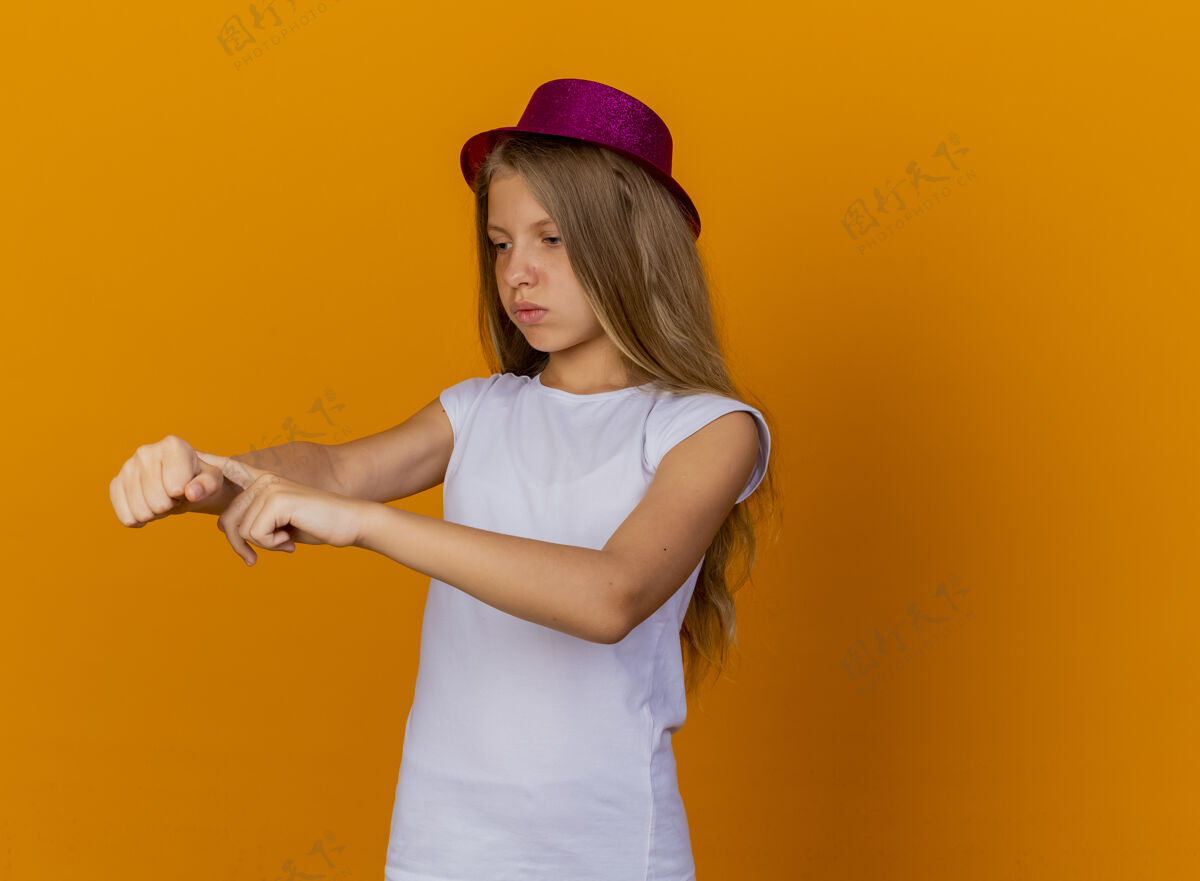 漂亮戴着节日帽子的漂亮小女孩指着她的手 提醒着时间不满意 生日聚会的概念手帽子提醒