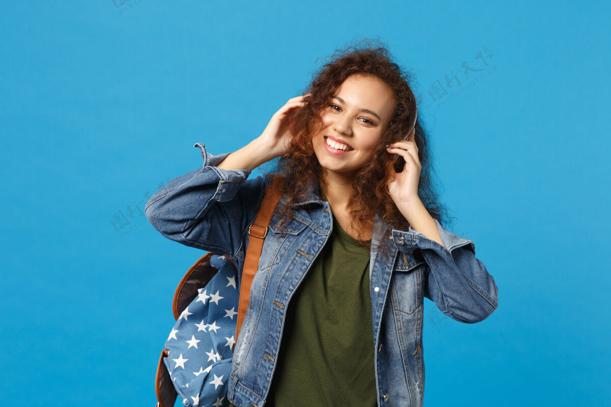 女人年轻的非洲裔美国少女 穿着牛仔服 背着耳机 隔离在蓝色的墙上包享受音频