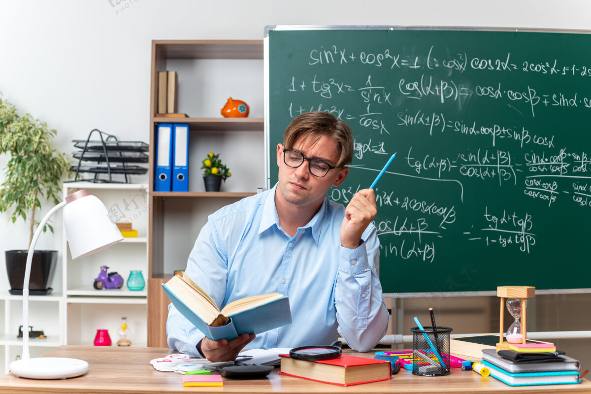 严肃年轻的男老师戴着眼镜坐在书桌旁 拿着书和笔记 严肃的脸在教室的黑板前看书阅读黑板课桌