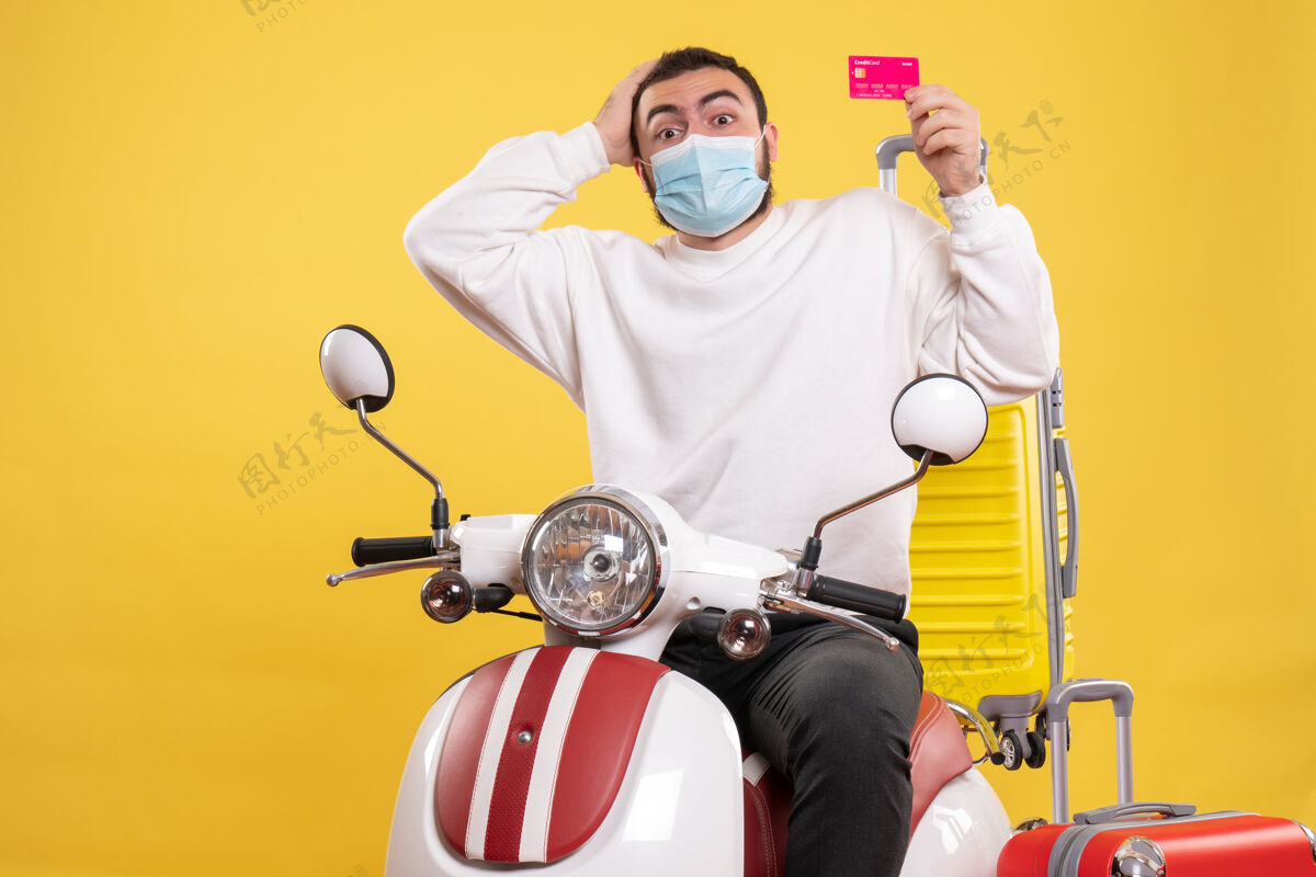 男旅行概念的俯视图 戴着医用面罩的困惑的家伙坐在摩托车上 黄色手提箱在上面 手里拿着银行卡银行黄色持有