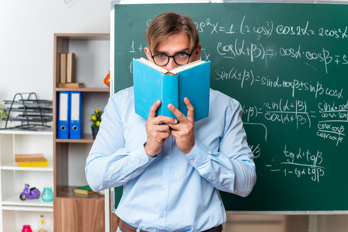 严肃年轻的男老师戴着眼镜拿着一本严肃的书站在教室里数学公式的黑板旁黑板公式年轻