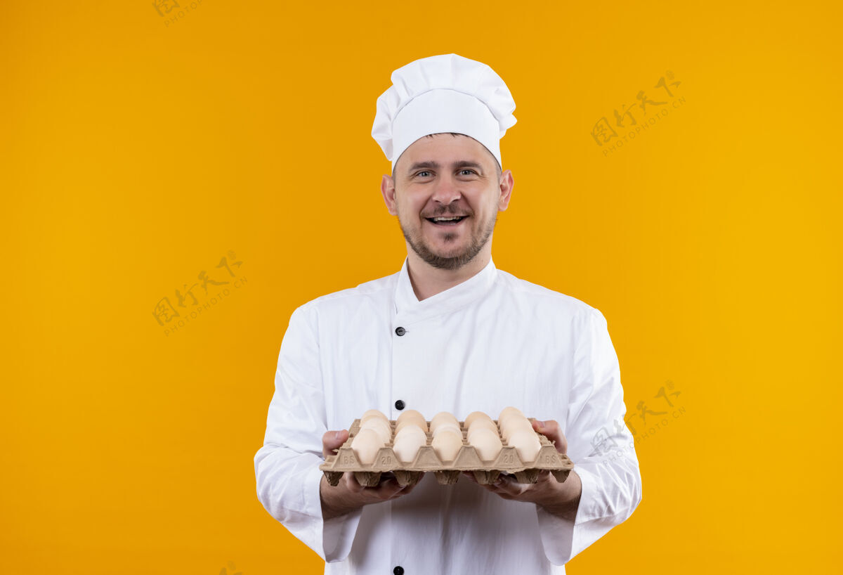 帅气快乐的年轻帅哥厨师身着厨师制服 手里拿着一盒孤立在橙色墙上的鸡蛋年轻持有纸箱