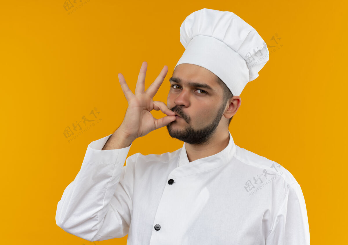 手势自信的年轻男厨师身着厨师制服 在橙色的墙上做着美味的手势男性制服自信